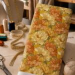 Oversized ivory and burnt orange blooms with muted olive foliage over a rich mustard-yellow background.